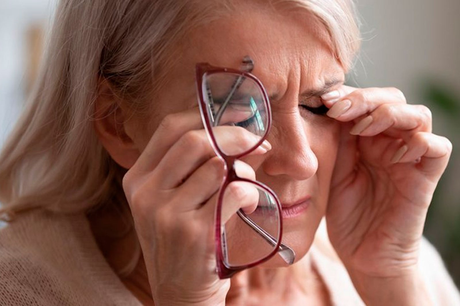 Mulher madura expressando desconforto visual, segurando seus óculos de grau e esfregando os olhos com cansaço. Imagem ilustrativa para a seção de Cirurgia Facorrefrativa com implante de lentes intraoculares premium realizada pela Dra. Andressa Guimarães na Clínica de Olhos da Cidade, Rio de Janeiro.